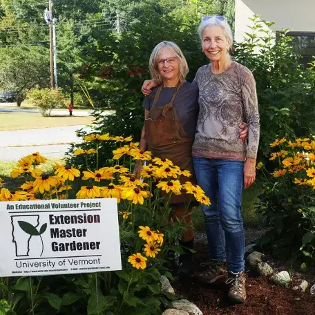 2 people smiling with their arms around each other, standing outdoors next to a group of flowers and a master gardener sign