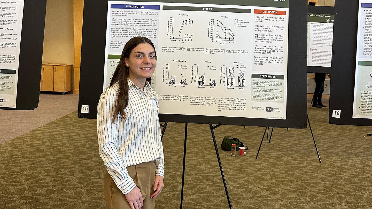 young woman with long brown hair and white shirt standing in front of research poster at conference event