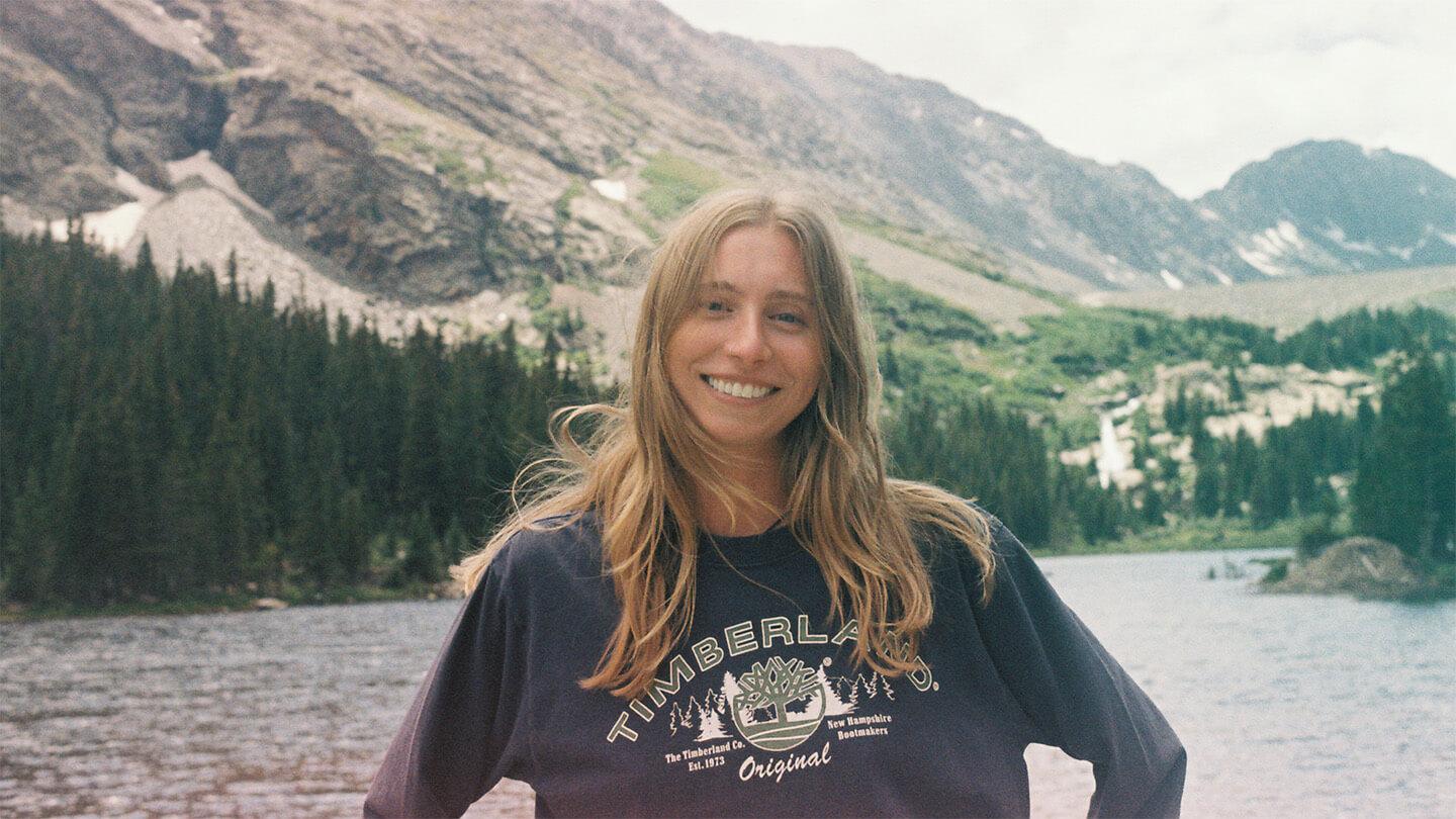 young woman with long, light hair standing in front of mountain and lake