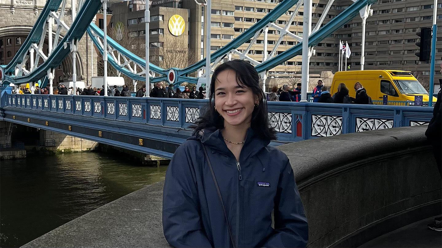 young Asian person with long dark hair wearing blue jacket standing in front of bridge in urban setting