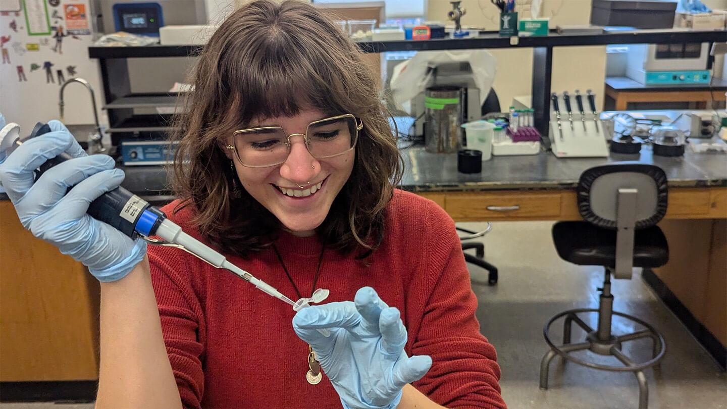 young woman with long brown hair, red shirt, and safety glasses wearing blue gloves and injecting something in a lab