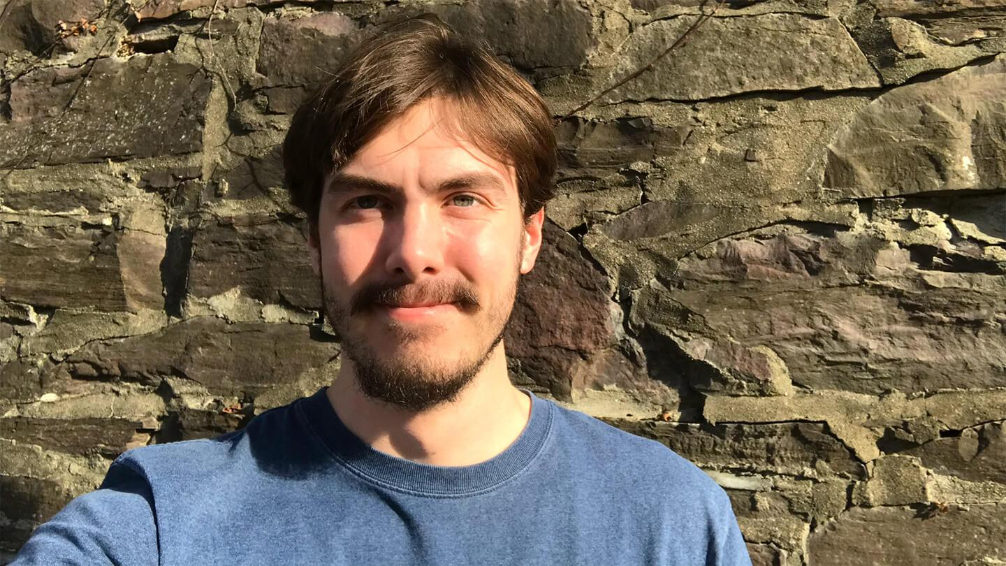 young man with brown hair, mustache, and beard, wearing blue t-shirt, taking selfie in front of stone wall