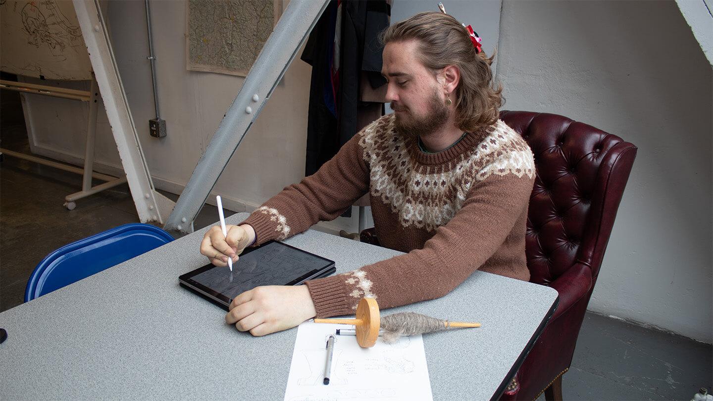young man with brown hair and beard and brown sweater sitting at table drawing on tablet with paper drawing and pencil beside him