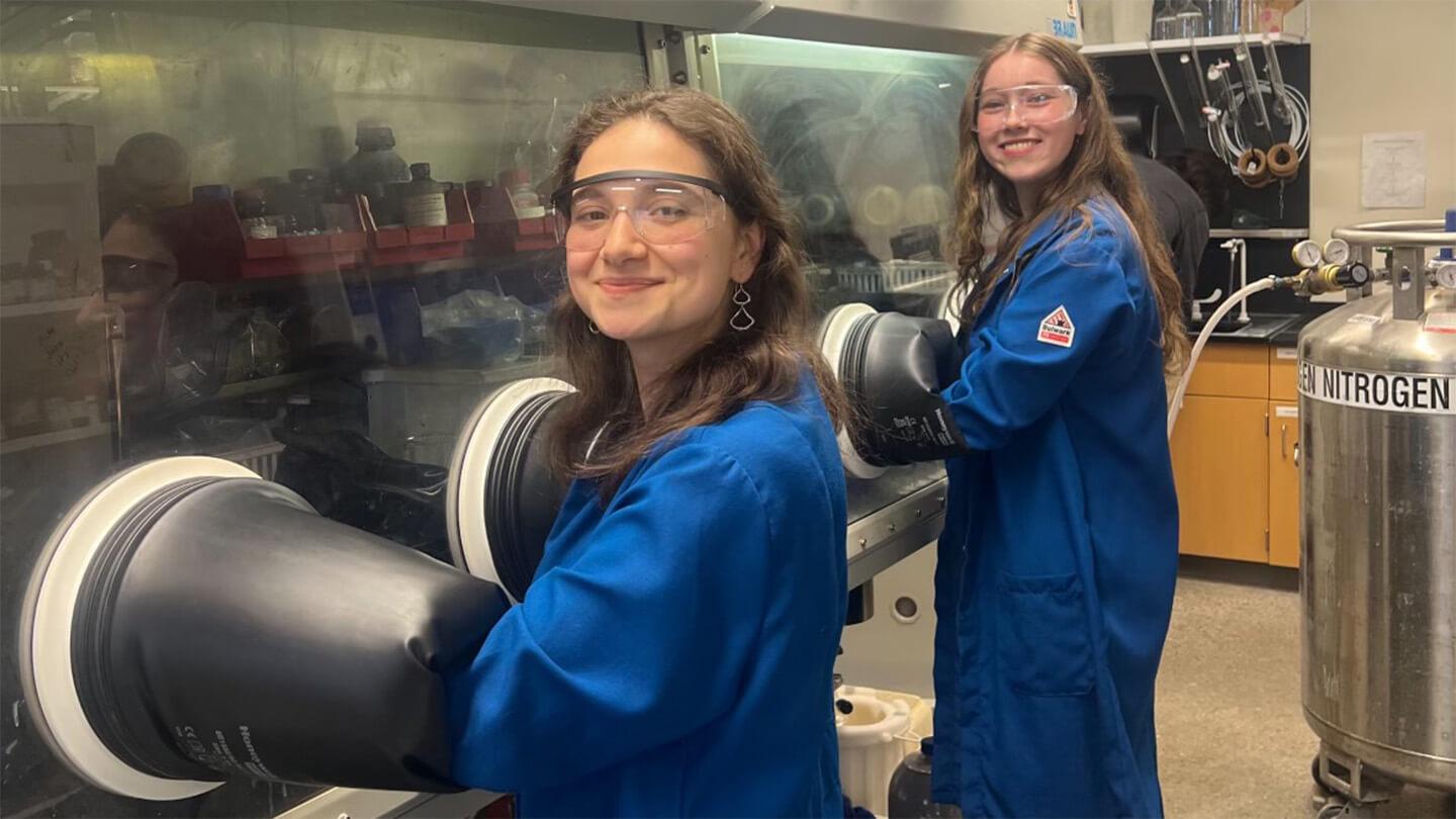 two young women in blue lab coats and safety glasses working in lab