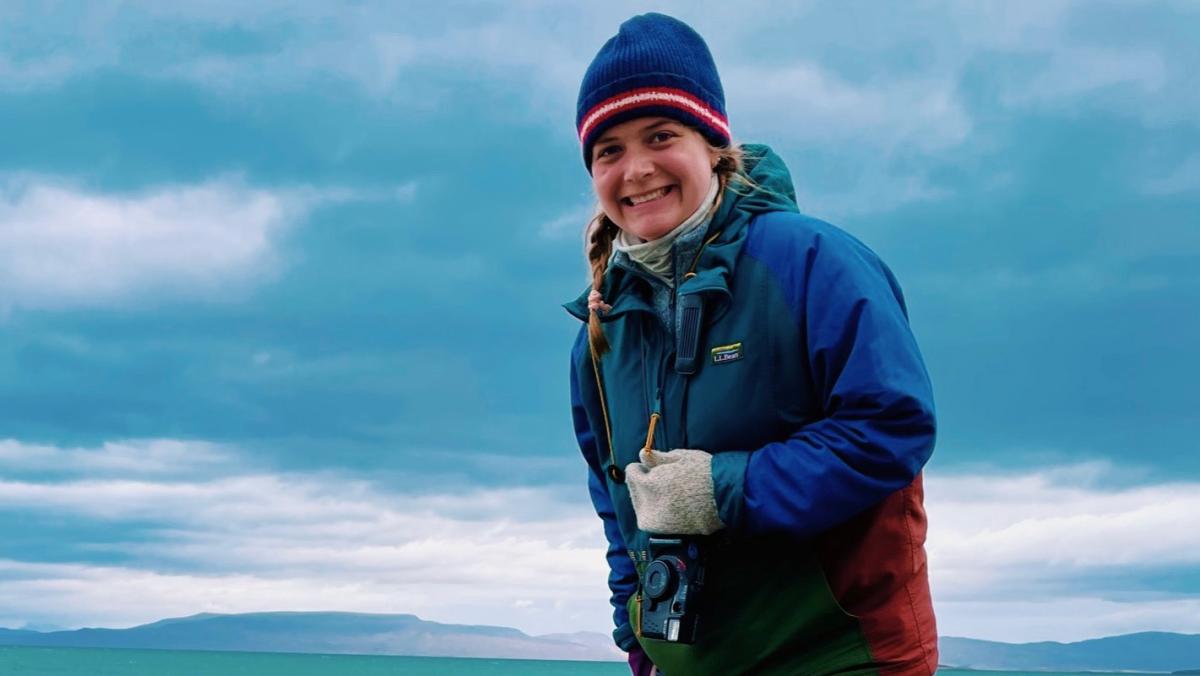 Smiling young woman in colorful jacket and hat with camera around neck with ocean in background