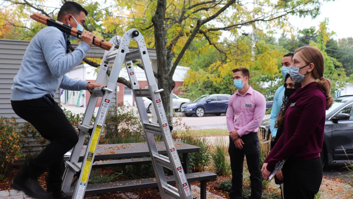 Person climbing a ladder while holding a long object over their shoulder, and three other people watching.