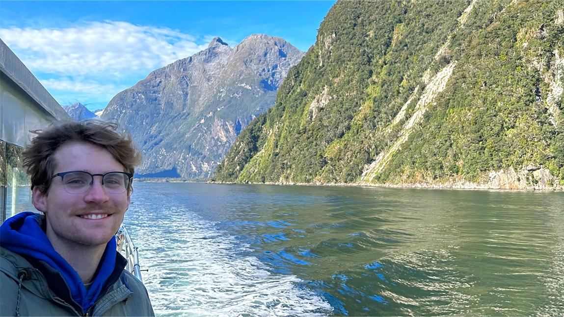 Cole on the side of a boat with water and mountains behind him.