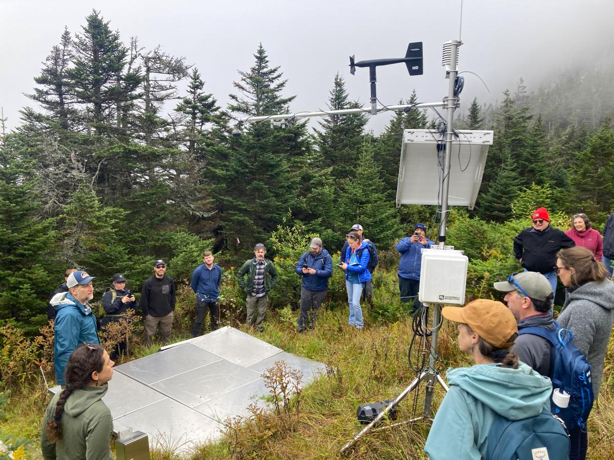 Dr. Arne Bomblies introduces the Summit-to-Shore Snow Monitoring Network in Vermont to a group of researchers. They crowd around a snow monitoring system atop Mount Mansfield. 