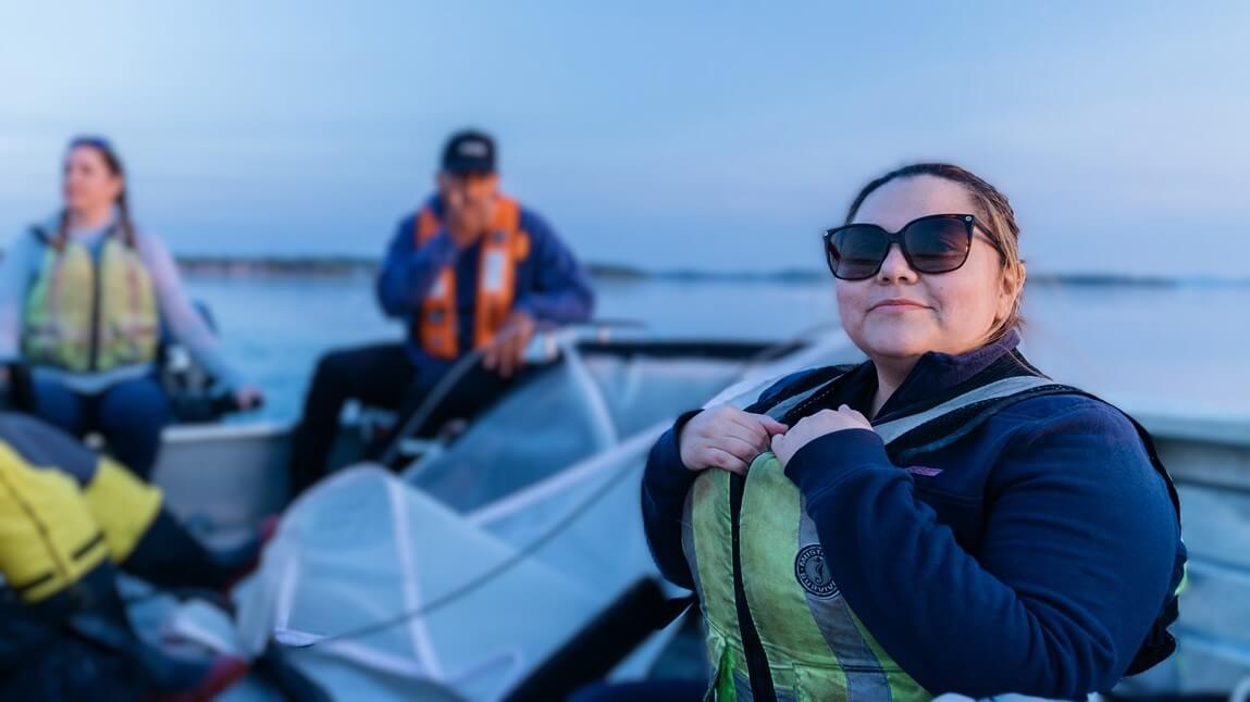 Rosie Chapina sitting on a boat, two people are in the background, all are wearing life jackets. Rosie is smiling and has sunglasses on.