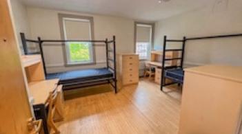 A room with wood floors and two beds, two dressers, and two desks. 