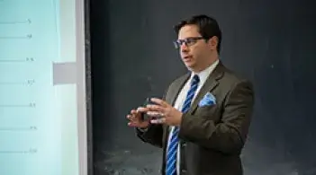 A man in a suit and tie standing in front of a blackboard giving a lecture