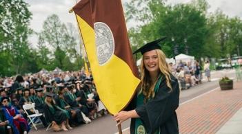 A student at UVM Commencement holding the Rubenstein School flag and wearing a cap and gown