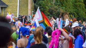 A large and diverse group of community members walking through downtown Burlington as part of a pride parade. An inclusive LGBTQ+ flag is in focus among the sea of people.