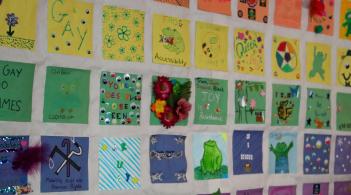 Squares of paper in orange, yellow, green, blue, and purple lined up and taped to a wall in neat rows. Each square has a drawing, phrase, or collage crafted onto it with LGBTQ+ and social justice themes, aspirations, and memes. 