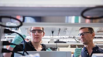 student conducts research in a lab while professor oversees.