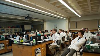 A group of individuals in lab coats seated at desks, engaged in collaborative work within a laboratory setting