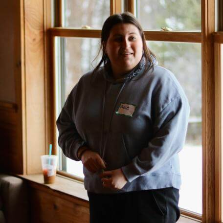 A UVM student engages in animated discussion during Social Justice Grounding Retreat. She is standing in front of the wood windowsill of Common Ground retreat center, with snow covering the ground outside behind her. 