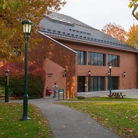 A brick building with large windows. There are trees and green grass and a path in the foreground.