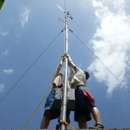 Two people securing equipment to a wind measurement tower under a blue sky.