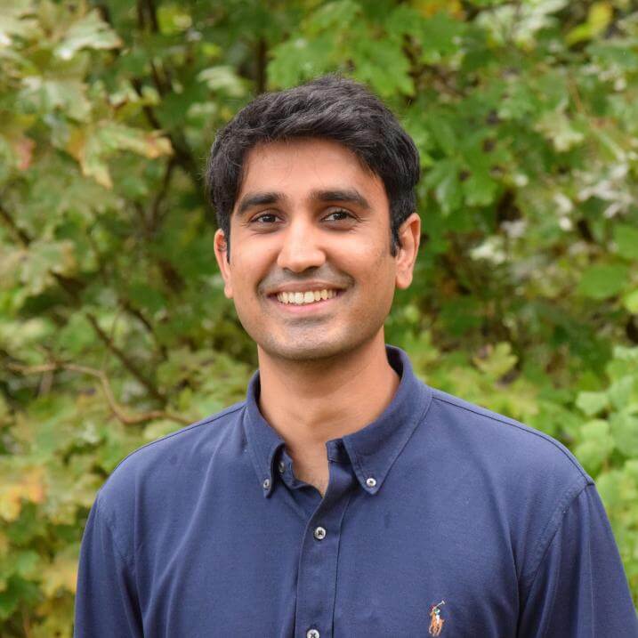 Ali Mahmood standing and smiling in a navy blue button down shirt in front of greenery