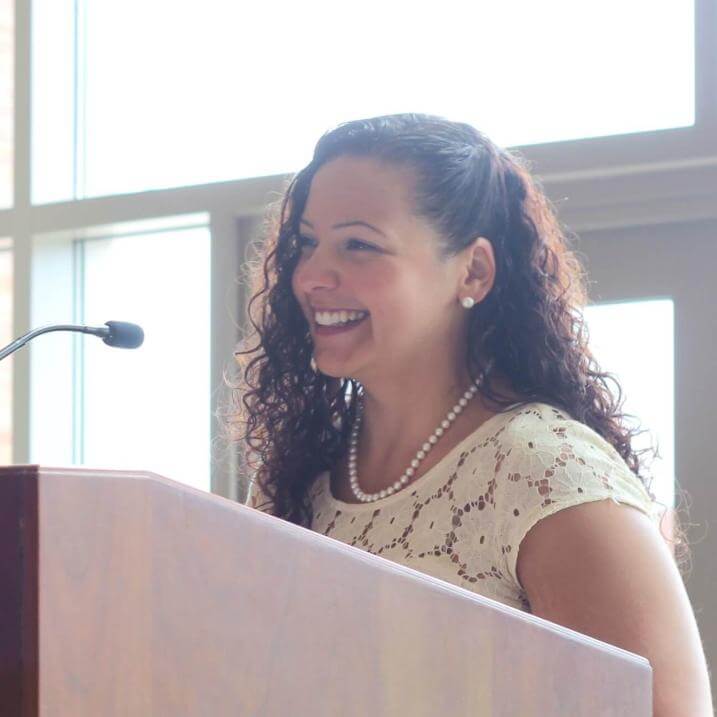 Jaydeen Santos standing and smiling at a podium. She is wearing a white eyelet dress.