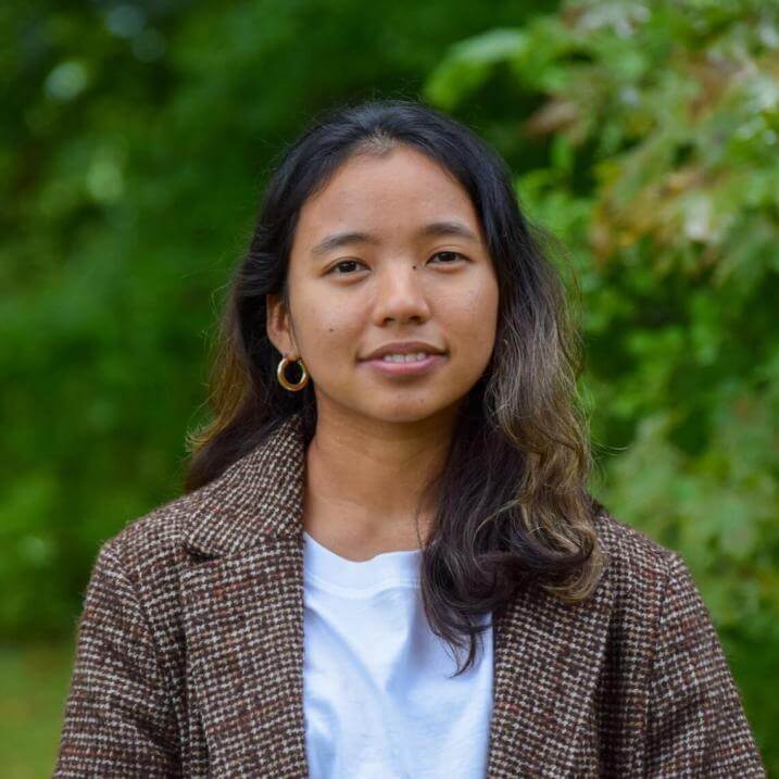 Menuka standing in a tweed jacked and white t-shirt in front of greenery