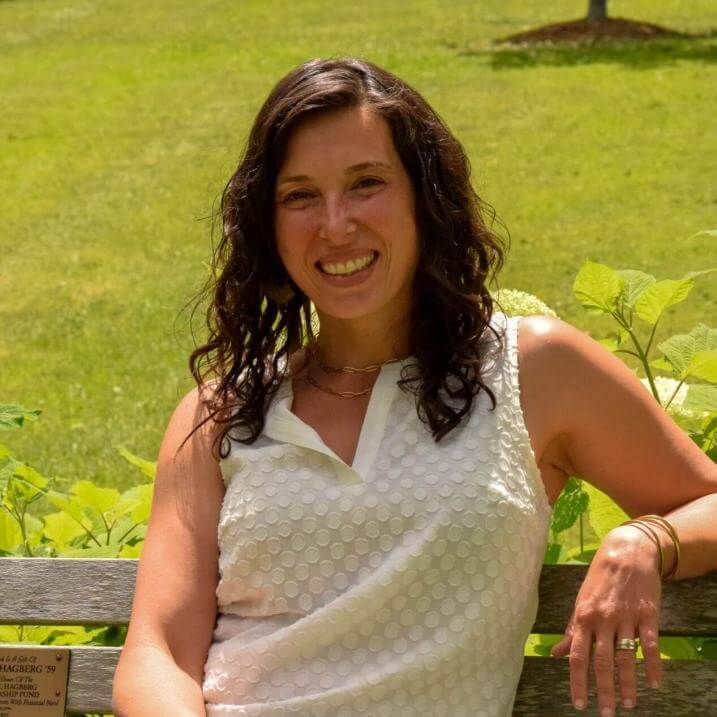 Sandy sitting on a bench outside with a green lawn behind her. She is wearing a white tank top and smiling.