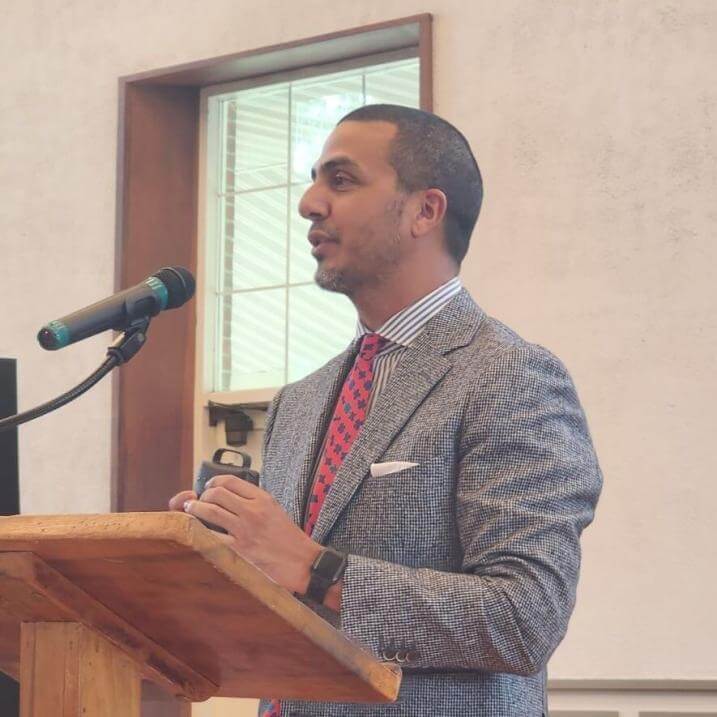 ahmed khanani standing at a podium talking. they are wearing a tweed jacket and red patterned tie.