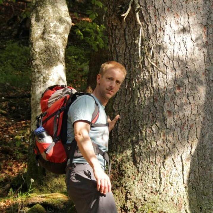 Bill Keeton standing next to a large tree in a forest wearing a red backpack