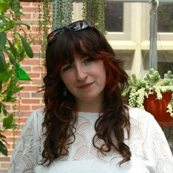 Allie stands in front of multiple hanging plants. She has red highlights in her hair and is wearing a white shirt.