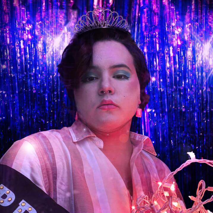 Juneau stands wearing a prom sash and holding string lights in front of a purple background. They wear a crown and green eyshadow in a pink and white striped shirt.