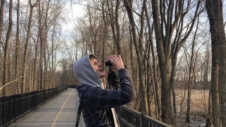 Pierre standing on a path in the woods looking up with a pair of binoculars.