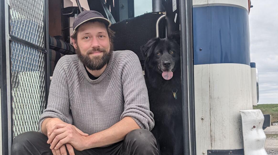 Serge Wiltshire. A man with dark hair and a beard in a grey sweater and hat sits with a black dog. 