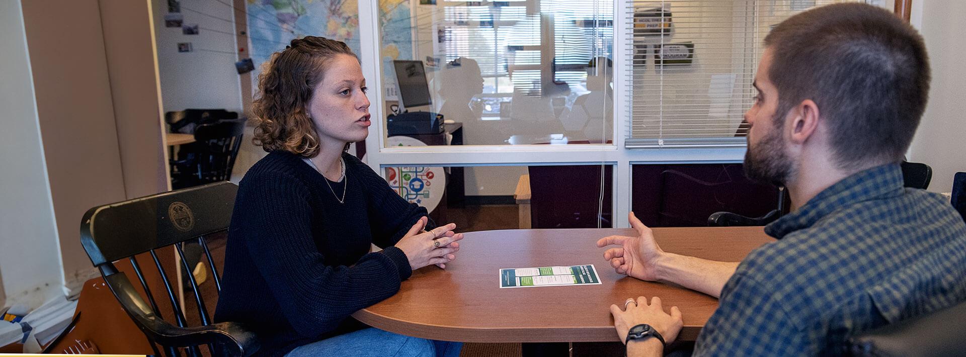 two individuals talking at a desk.