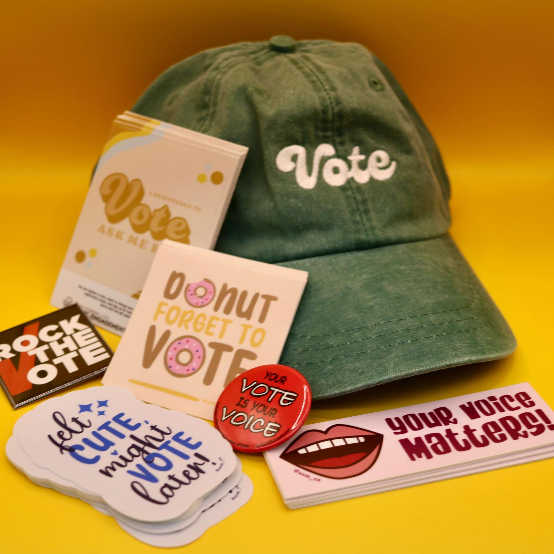 A collection of stickers, buttons and hats designed as part of Civic Engagement's Voter Education campaign, displayed in front of a yellow background