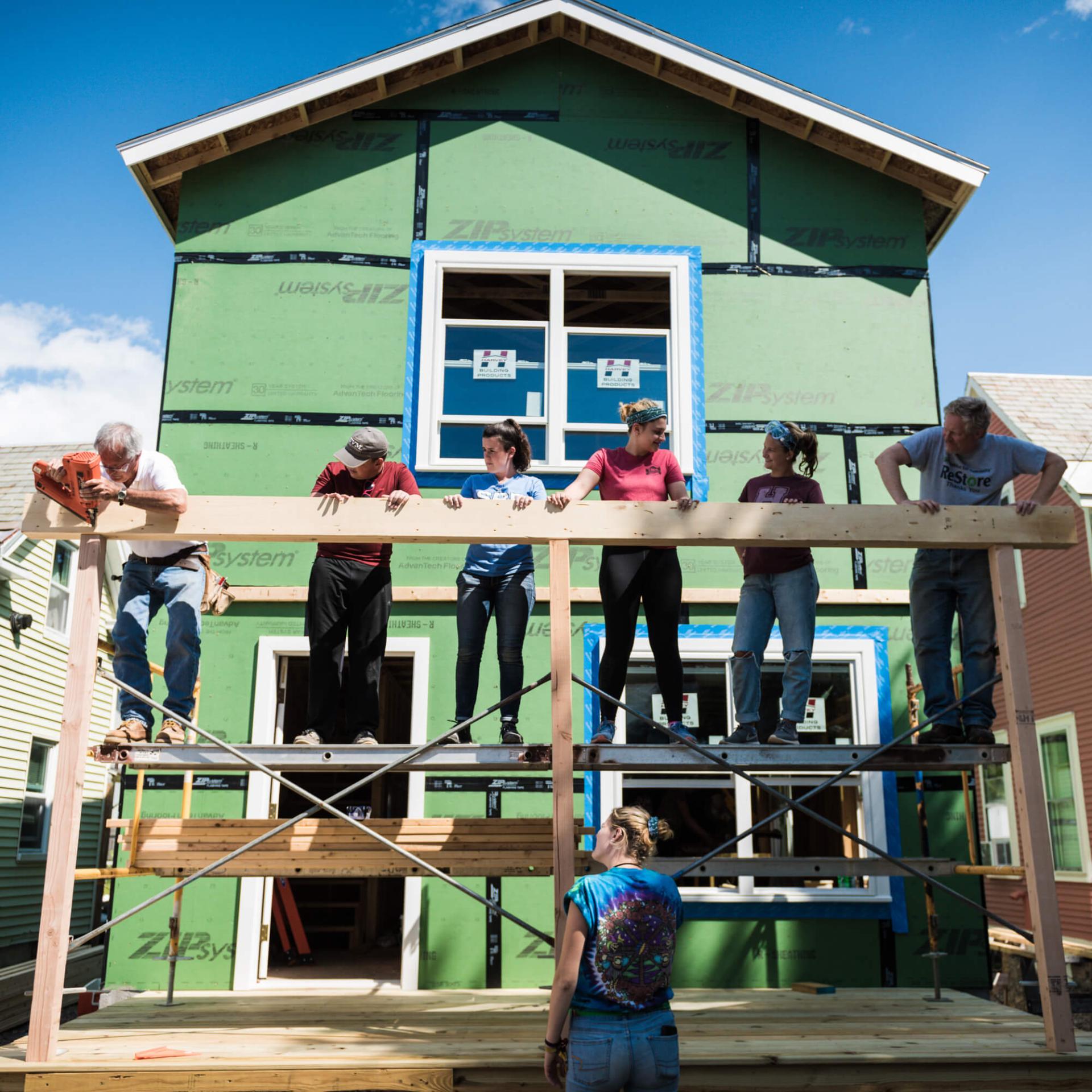 A group of participants work together to balance a beam in front of a large Habitat for Humanity build