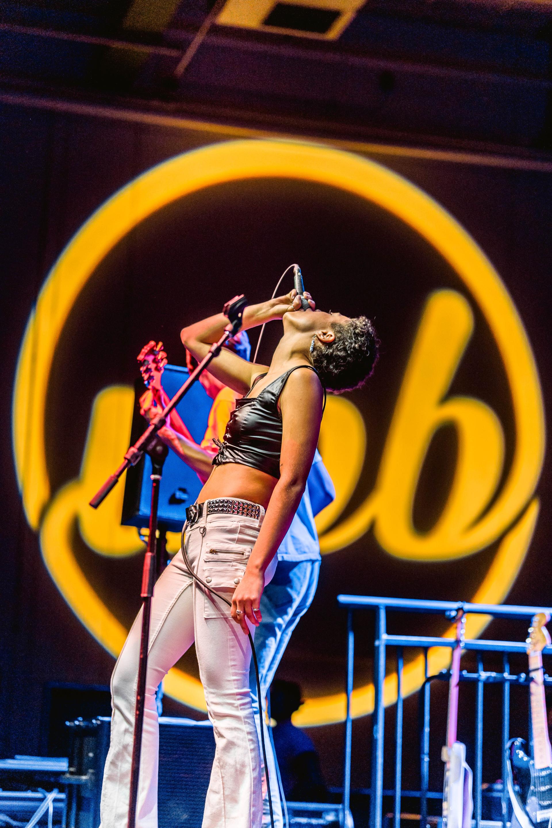 Photo of a singer leaning her head and body backwards while singing passionately into a microphone, with a large yellow glowing University Program Board logo displaying on the wall behind her at FallFest