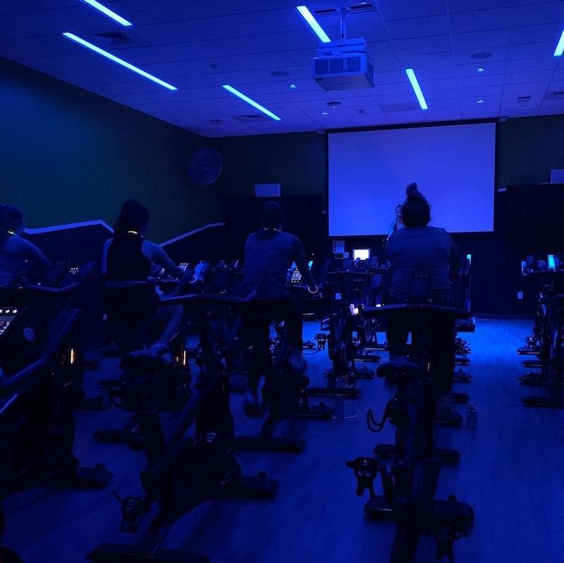 Students on stationary bikes in a black-lit group fitness class. 