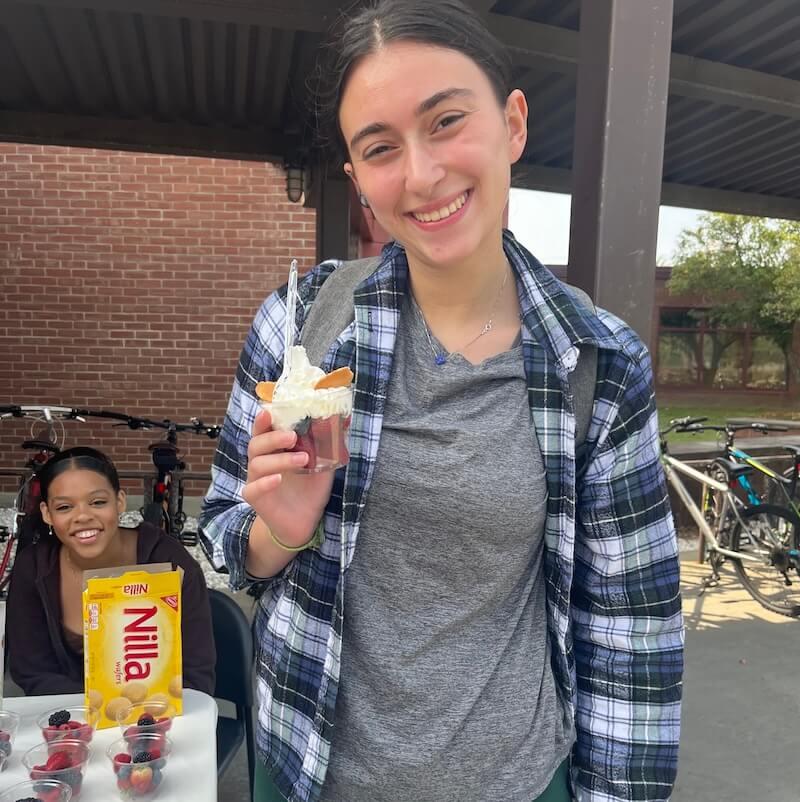 A student standing next to a table outside holding a sundae. 