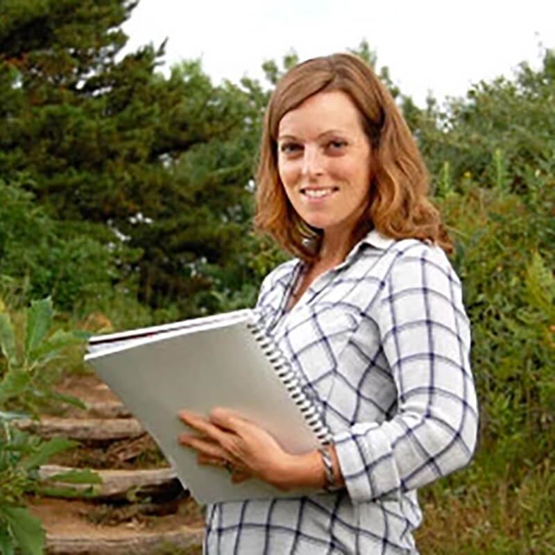 A student holding a notebook, standing on a path, surrounded by nature