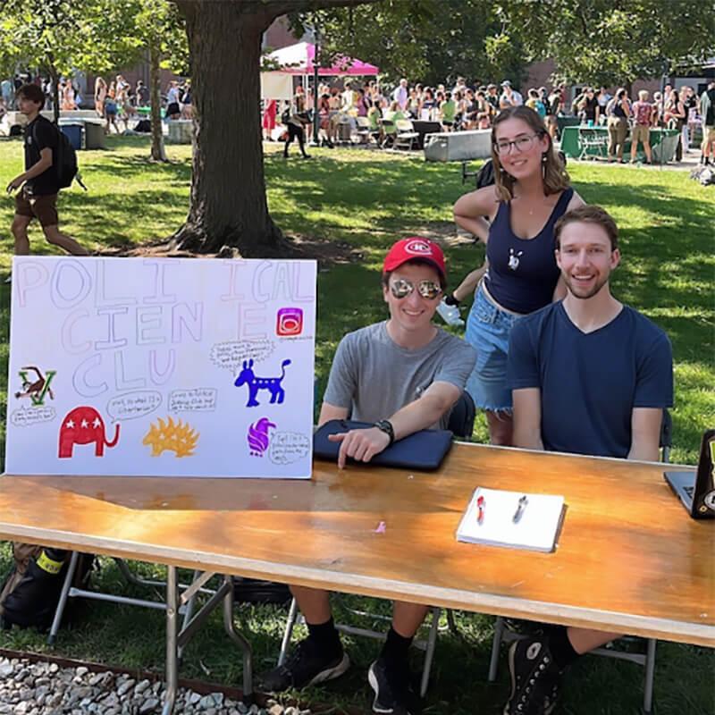 Three people sitting at a table with a political science club sign