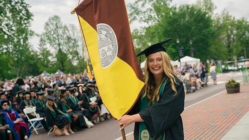A student at UVM Commencement holding the Rubenstein School flag and wearing a cap and gown