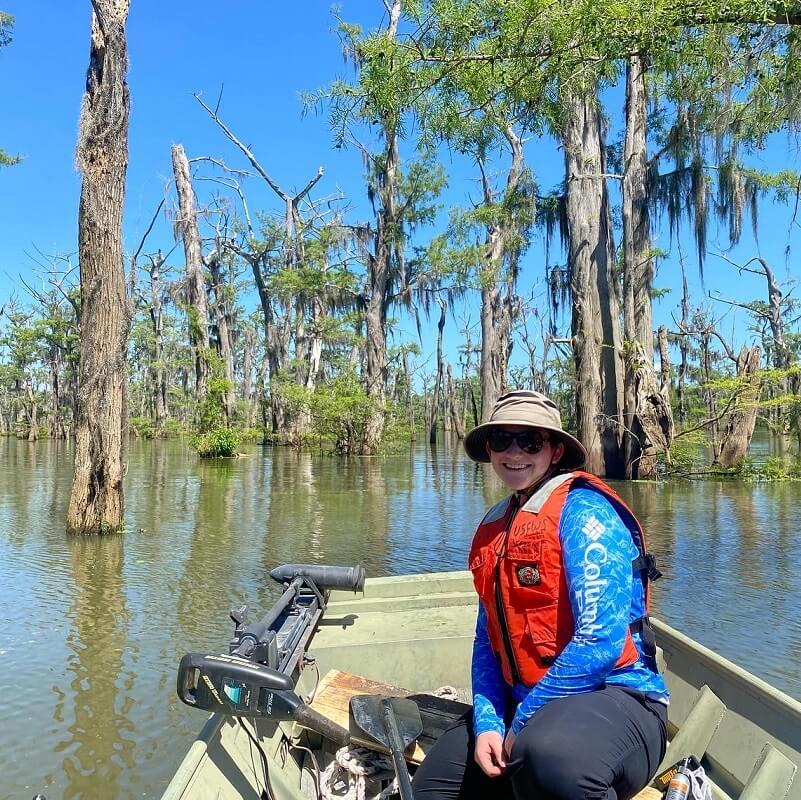 A student sitting in a small boat by themselves wearing a life jacket, hat, and sunglasses. They are smiling and there are wetland forest behind them.