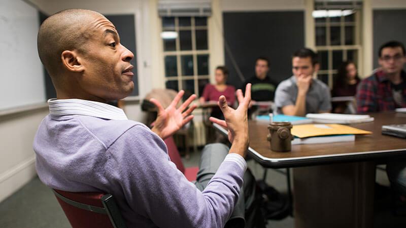 a professor sits in a chair gesturing with his hands while a class looks on