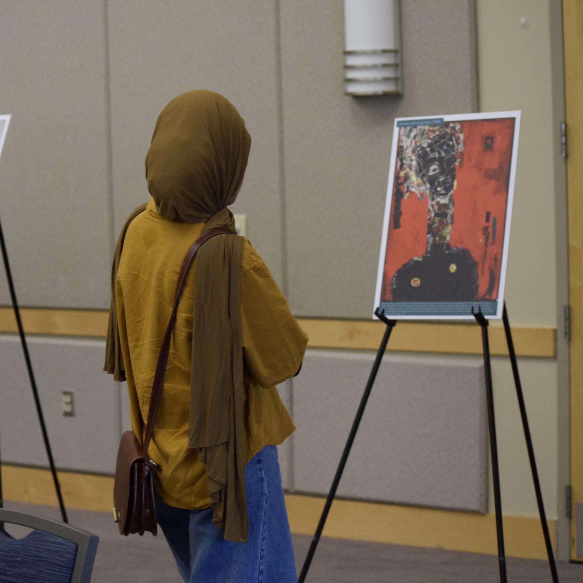 Woman wearing a hijab facing away from the camera looks at a print of artwork on an easel. 