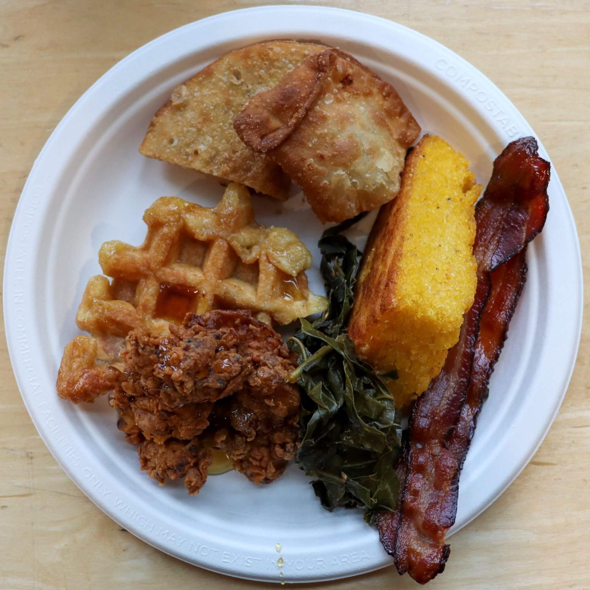 Plate of breakfast food including bacon, cornbread, collard greens, empanadas, fried chicken, and a waffle