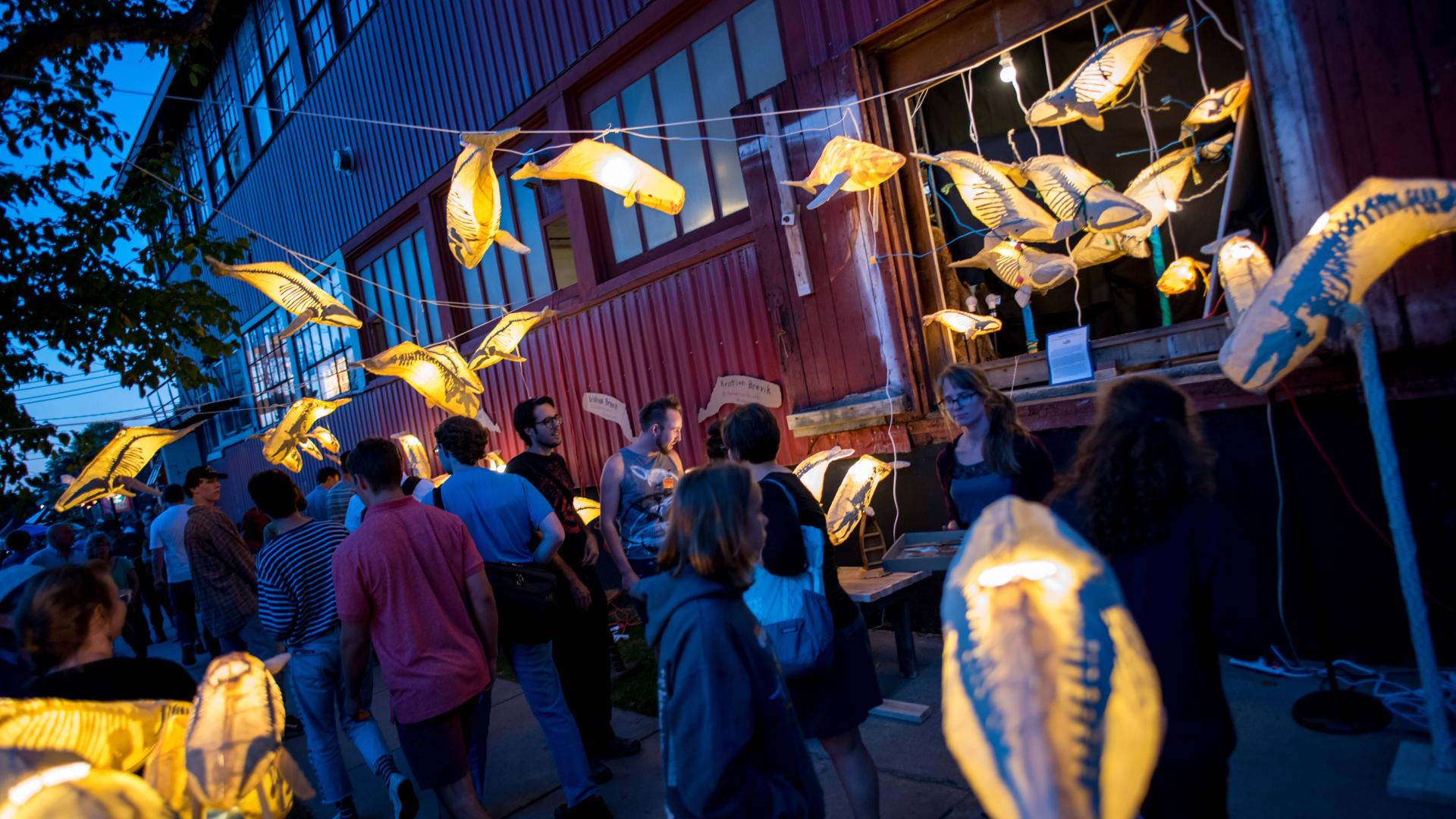 Downtown Burlington at twilight with a crowd of people walking in front of a warehouse. Paper lanterns shaped like whales are strung up in the foreground.