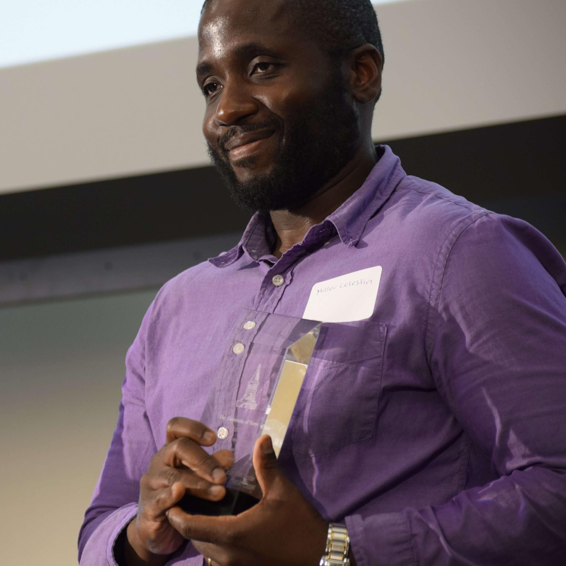 Miller Celestin wearing a purple button down shirt and smiling as he holds an acrylic award.