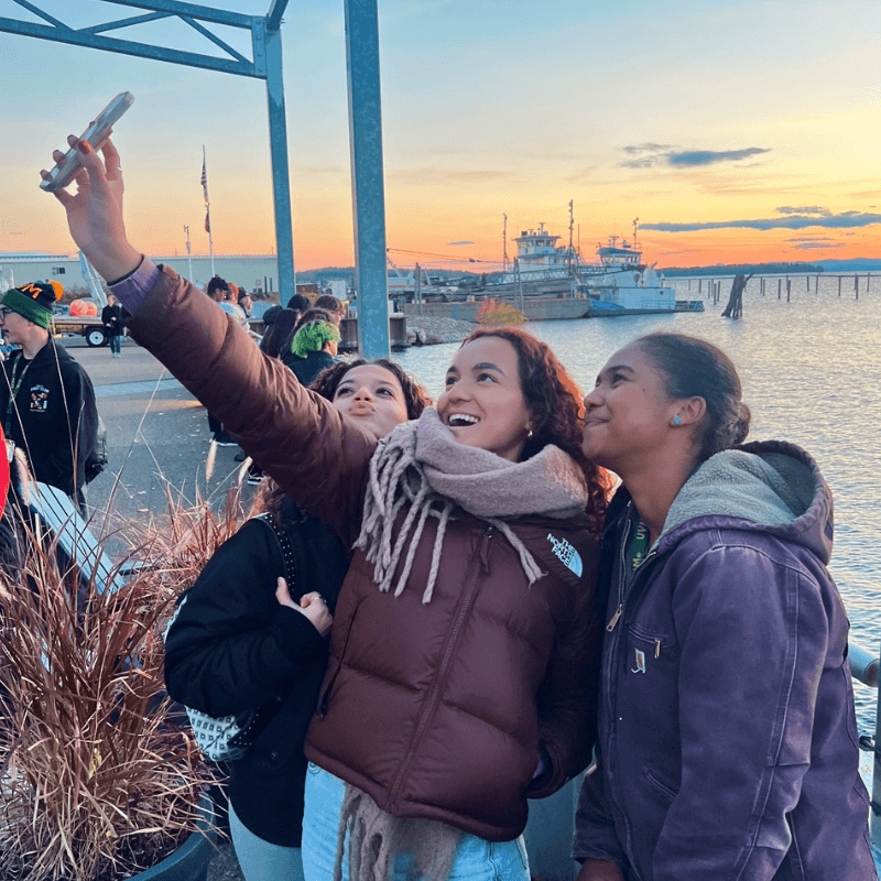 Three students take a selfie in front of Lake Champlain at sunset