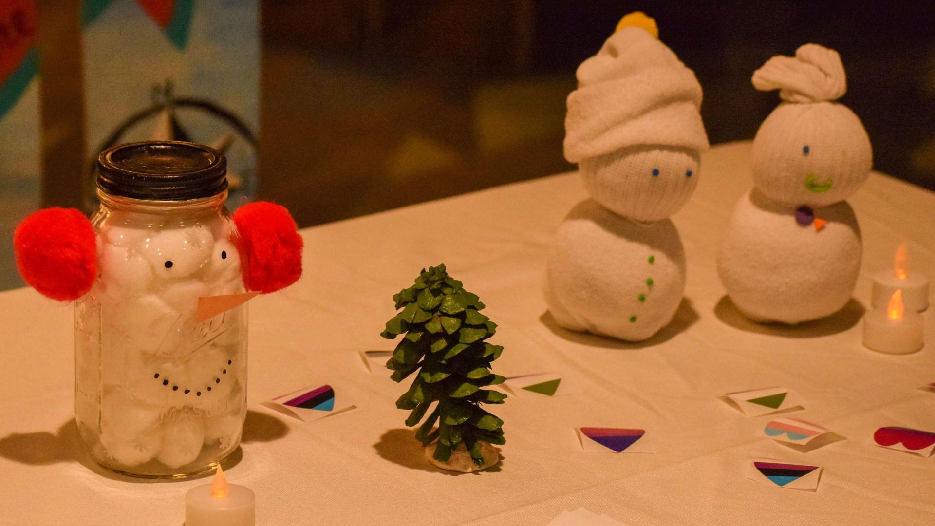 Three crafted snow people, one made from a mason jar filled with cotton balls, and two made from socks, sit on a table peppered with candles, pinecones, and Prism Center sticker.